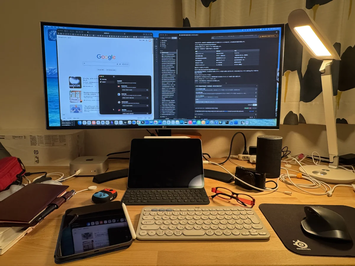 夜のデスク。ウルトラワイドモニターにブラウザとAIツールの画面を並べ、手前にMac mini・iPad・キーボード・マウスが並んでいる
