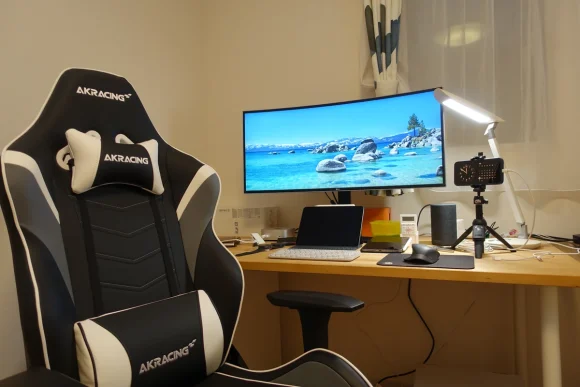 白い部屋の作業デスクを斜めから見た全景。手前に白と黒のAKRacingのゲーミングチェア、奥の木製デスクにLGの34インチ曲面ウルトラワイドモニターとデスクライトが並んでいる