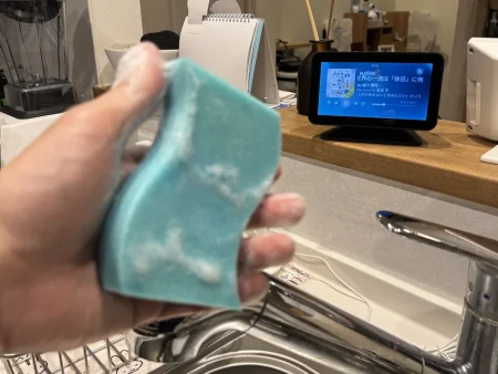 皿洗いの手元。泡のついたスポンジを持つ手の奥で、キッチンカウンターのEcho Show 5がオーディブルの本を再生している