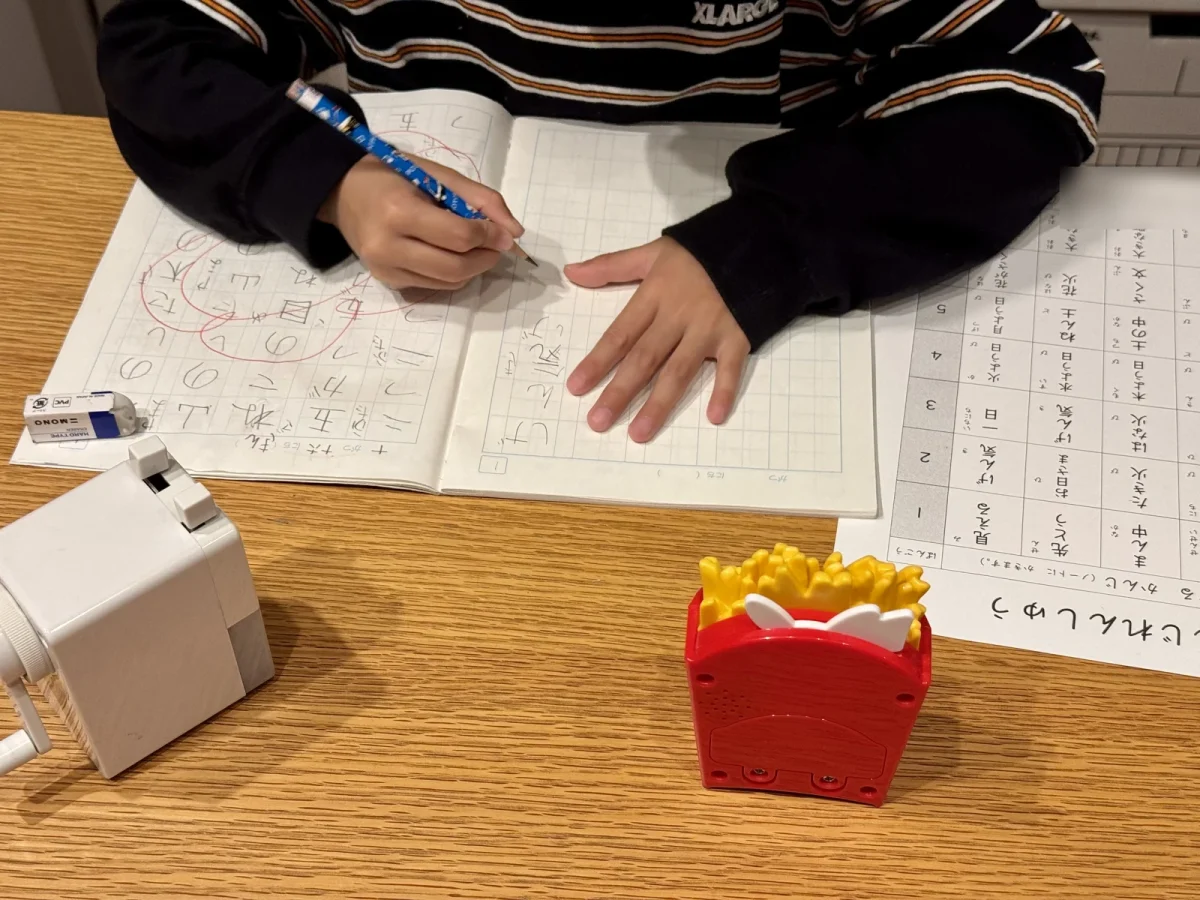 ダイニングテーブルで漢字ノートに書いている小1の手元と、手前に置いたフライドポテトの形の赤いキッチンタイマー